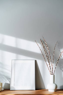 Modern minimalist style interior with white poster mockup, candles and blooming branches of the pussy willow in vase on wooden console under sunlight and shadows on a white gray wall. Selective focus