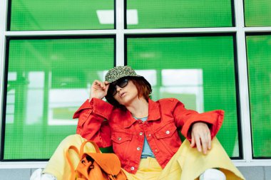 Parlak elbiseli, güneş gözlüklü, sırt çantalı ve kova şapkalı genç bayan yeşil cam arka planda otururken poz veriyor. Şehir sokak modası. Moda blogcusu. Seçici odak