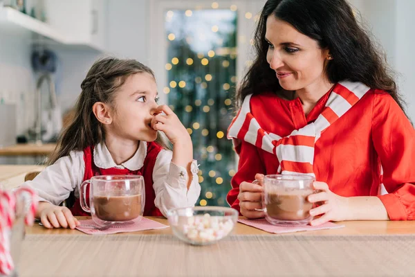 サンタの帽子の幸せな笑顔の娘の女の子と彼女のお母さんは 装飾されたモダンなキッチンに座って マシュマロでココアを飲んでいます 家族の休日だ クリスマスのお祭り気分 選択的焦点 — ストック写真