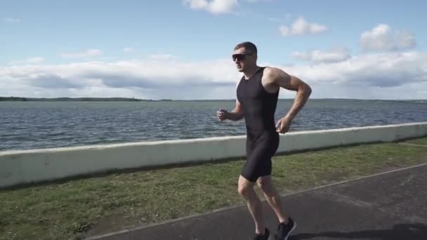 Corredor profesional corre en una carretera cerca del lago, atleta entrena en un día soleado, corriendo antes de las competiciones de triatlón. — Vídeos de Stock