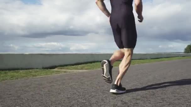Corredor corre en una carretera cerca del lago, atleta entrena en un día soleado, vista de bajo ángulo, competiciones de triatlón. — Vídeos de Stock