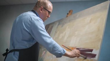 Sanat stüdyosu, mavi gömlekli ve gözlüklü yaratıcı kişi, yetenekli yetişkin bir erkek sanatçı, büyük bir tuvale soyut bir resim çizer..
