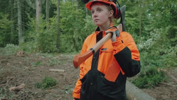 Vrouwelijke houthakker staat in het bos, een jonge gespecialiseerde vrouw in beschermende kleding houdt een bijl in haar handen en werkt aan ontbossing. — Stockvideo