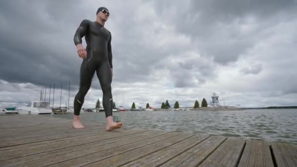 Nadador profesional, triatleta en traje de neopreno camina a lo largo del muelle y se prepara para saltar al agua, 4k cámara lenta. — Vídeo de stock
