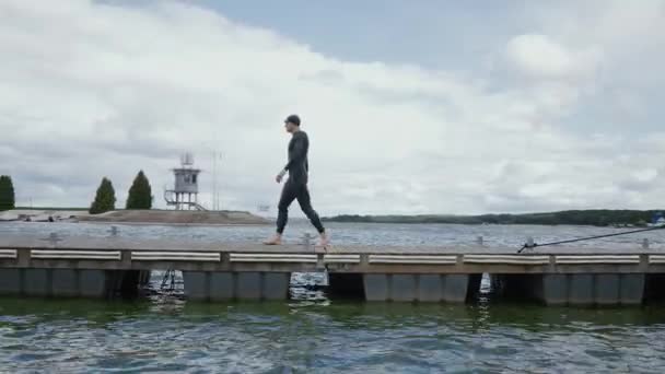 Nadador profesional en traje de neopreno camina a lo largo del muelle y salta a aguas abiertas, un atleta entrena antes de una competición de triatlón. — Vídeos de Stock