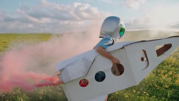 Dreamer boy juega en un casco de astronauta en la naturaleza con un modelo de cartón de un transbordador espacial, un niño corre a través de un campo de colza soñando con volar al espacio, humo rosa, 4k cámara lenta. — Vídeos de Stock