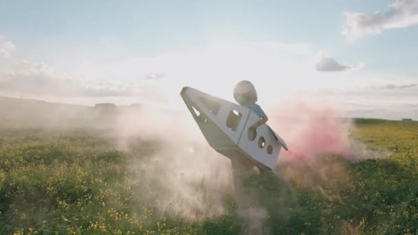 Campo, niño juega en un casco de astronauta en la naturaleza con un modelo de cartón de un transbordador espacial, un niño corre a través de un campo de colza soñando con volar al espacio, humo rosa, 4k cámara lenta. — Vídeos de Stock