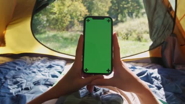 De mano, vista en primera persona, mujer sosteniendo un teléfono inteligente con una pantalla verde mientras está acostado en una tienda de campaña, croma plantilla clave, recreación al aire libre. — Vídeos de Stock