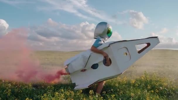Хлопчик грає в астронавтському шоломі на природі з картонною моделлю космічного човника, хлопчик проходить через поле ріпаку, що мріє про політ у космос, рожевий дим, повільний рух 4k . — стокове відео