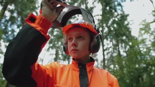 Retrato de una joven maderera parada en el bosque, especialista en equipo de protección abre la visera del casco de protección y mira a la cámara, 4k cámara lenta. — Vídeo de stock