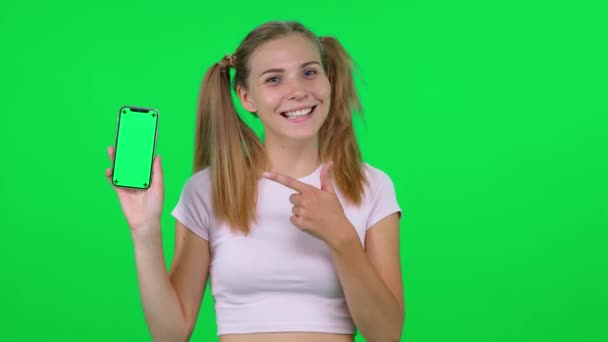 陽気な白人女性は彼女の手にスマートフォンを持っており、カメラにそれを示しています,若い女性は彼女の感情を共有し、彼女のスマートフォンを示しています, chromakeyテンプレート. — ストック動画