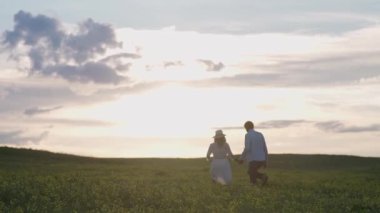Kırsal kesimde, doğada rahatlayan bir çift neşeli insan, bir erkek ve bir kadın kolza tohumu tarlasında koşuyorlar, el ele tutuşuyorlar, gün batımı, bulanık görüş.