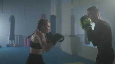 Kadın boksör yumruklarını ve savunmasını eğitiyor, odaklanma eldivenlerini yumrukluyor, boks salonunda bir erkek koçla antrenman yapıyor, 4 km yavaş çekim..