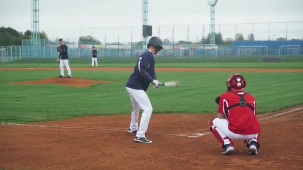 Студенческие виды спорта, парни играют в бейсбол, питчер бросает мяч в сторону отбивающего, отбивающий успешно бьет быстрый мяч и бежит на базу. — стоковое видео