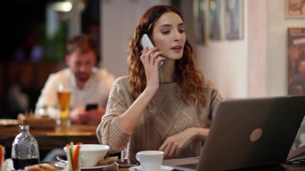 Młoda kobieta freelancer pracuje poza domem, kobieta deweloper siedzi w barze i pracuje w laptopie, rozmawiając przez telefon komórkowy. — Wideo stockowe