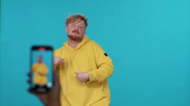 Sarı kapüşonlu genç, sosyal ağlar için akıllı telefondan video çekerken mavi ışık arka planında dans ediyor..