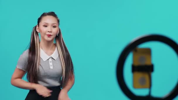 Asyalı bir kadın mavi ışık arka planında müzik eşliğinde dans ediyor ve sosyal bir ağ için akıllı telefondan kendini videoya çekiyor.. — Stok video