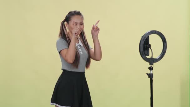 Genç Asyalı kadın bej arka planda müzik eşliğinde dans ediyor ve sosyal bir ağ için akıllı telefondan kendini videoya çekiyor.. — Stok video