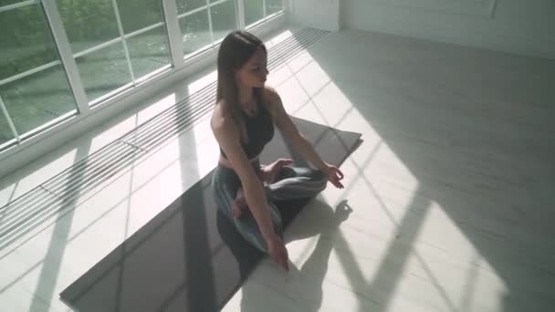 Wellness, jonge vrouw mediteert en doet yoga, een rustige stemming, ontspannen in een witte kamer gevuld met licht, het meisje zit op de vloer, camera beweging. — Stockvideo