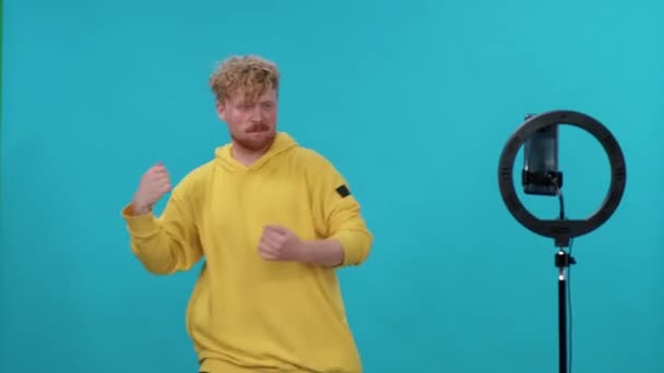 Ung man influencer i gul hoodie danser på en blå bakgrund och tar en selfie video på smartphone för ett socialt nätverk. — Stockvideo