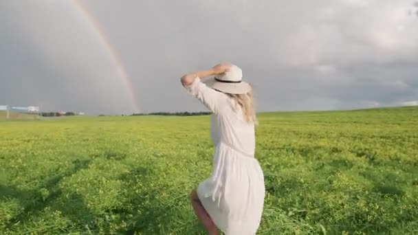 Countryside, baksidan av en glad ung flicka i vit klänning löper genom ett fält av raps, regnbåge efter regn, 4k slow motion. — Stockvideo