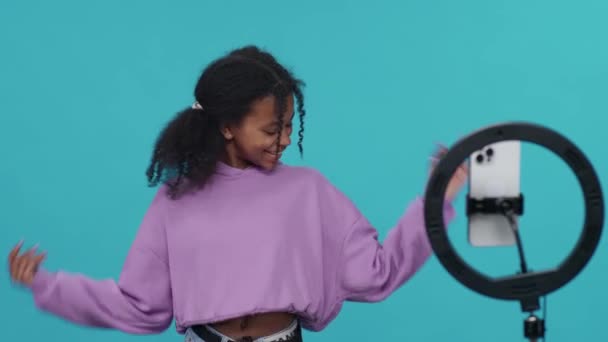 Genç Afrikalı bir kız mavi ışık arka planında dans ediyor ve sosyal ağ için akıllı telefondan bir selfie videosu çekiyor, kız hızlı hareket ediyor. — Stok video