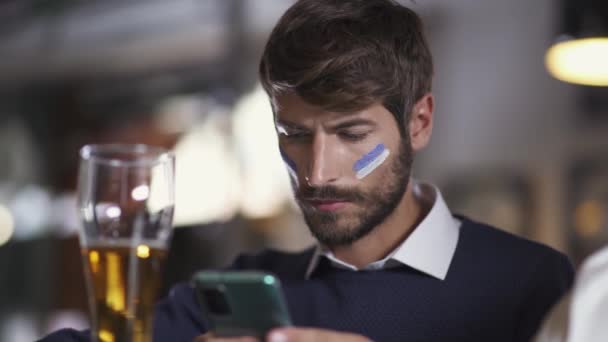 Fã de futebol senta-se em um bar durante um jogo de futebol, bebe cerveja e usando seu smartphone, navegar na internet. — Vídeo de Stock