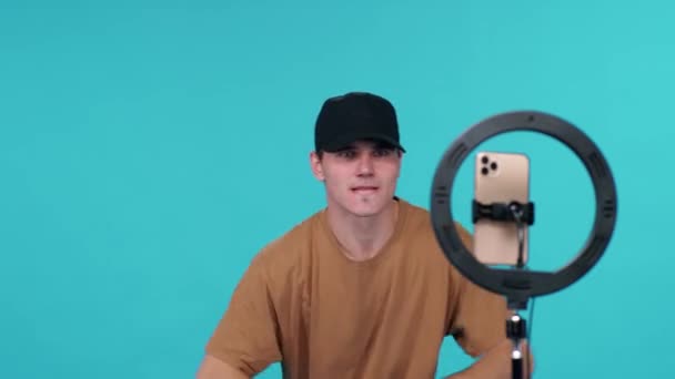 Young guy video blogger danst op de muziek op een blauwe achtergrond en neemt een selfie video op smartphone voor een sociaal netwerk. — Stockvideo