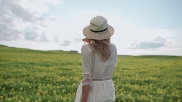 Platteland, achteraanzicht van een vrolijk jong meisje in witte jurk loopt door een veld van koolzaad, 4k slow motion. — Stockvideo