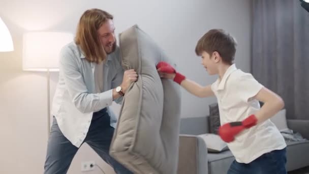Samoizolacja, ojciec i syn bawią się w boks, chłopiec w czerwonych rękawiczkach na rękach bije poduszkę na sofie, kwarantanna domowa podczas pandemii. — Wideo stockowe