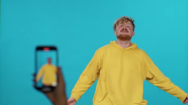 Sarı kapüşonlu adam mavi arka planda dans edip şarkı söylüyor ve sosyal ağ için akıllı telefondan bir selfie videosu çekiyor.. — Stok video