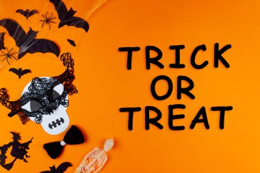 Kafatası canavarı maskesiyle komik bir yüz. Tuvalin sol tarafında karnaval malzemeleri var. Arka planda örümcekler ve yarasalarla parti aksesuarları. Siyah yazı, şeker mi şaka mı? Mutlu cadılar bayramı.