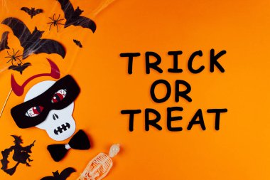 Kafatası canavarının maskeli ve boynuzlu komik yüzü. Tuvalin sol tarafında karnaval malzemeleri var. Arka planda örümcekler ve yarasalarla parti aksesuarları. Siyah yazı, şeker mi şaka mı? Mutlu cadılar bayramı.