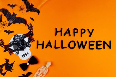 Kafatası canavarı maskesiyle komik bir yüz. Karnaval malzemeleri, tuvalin sol tarafında fotoğraf malzemeleri. Turuncu arka planda örümcekler ve yarasalarla parti aksesuarları. Siyah yazıt Mutlu Cadılar Bayramı.