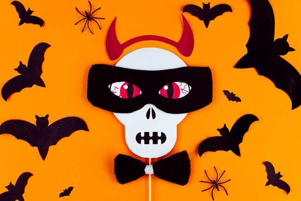 Funny face of skull monster with horns and mask made of photo props. Carnival supplies at center of canvas. Party accessories on orange background with black spiders and bats. Happy halloween.