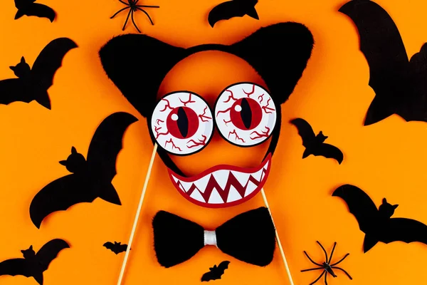 Funny face of monster made of photo props with headwear ears of cat. Carnival supplies at center of canvas. Party accessories on orange background with black spiders and bats. Happy halloween.