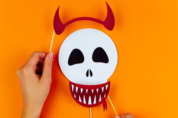 Funny face of skull monster with red horns and vampire smile on orange background. Female hands are holding paper photo props on canvas. Party accessories for celebration happy halloween.