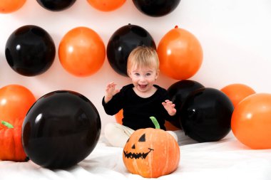 Cadılar Bayramı 'nı evde balkabağı ve balonlarla kutlayan beyaz bir bebek. Gülümseyen duygusal kız eğleniyor. 1 yaşındaki gülen çocuk, şenlikli dekorasyonun arasında kameraya bakıyor..