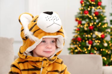 2022 yılının sembolü, kaplan kostümlü sevimli beyaz çocuk Noel ağacının arka planında oturuyor, gülümse, eğlen. Bebek tatilini evde kutla. Çocuk maskeli balosu. Yakın plan.