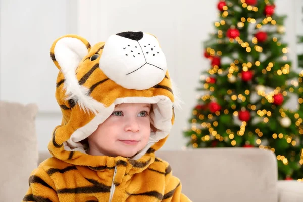 2022 yılının sembolü, kaplan kostümlü sevimli beyaz çocuk Noel ağacının arka planında oturuyor, gülümse, eğlen. Bebek tatilini evde kutla. Çocuk maskeli balosu. Yakın plan.