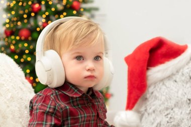 Noel ağacının yanında kulaklık takan sevimli sarışın, beyaz bir bebek. Ciddi bebek, Noel ve Yeni Yıl müziği, melodiler, kapsül kalıpları, kitap konsepti dinliyor. Boşluğu kopyala.