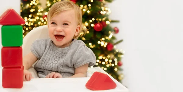 1-2 yaşlarındaki sevimli beyaz sarışın kız bebek Noel ağacının önünde oturuyor, gülüyor, gülüyor, gri arka planda parlak bloklarla oynuyor. Boşluğu kopyala..