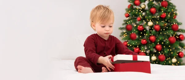 Mutlu, sarışın, beyaz bir bebek, evde beyaz bir battaniyeyle Noel ağacının yanında kırmızı bir hediye kutusu alıyor. Kış tatili süresince evde. Bebek, çocuk, pijamalı bir çocuğa yeni yıl hediyesi..