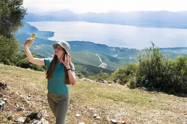 Güzel kız, kadın, dağlarda seyahat eden, görüntülü konuşma yapan, akıllı telefon görüşmeleri yapan, gülümseyen, el sallayan güzel göl kenarında. Yürüyüş sırasında iletişim. Trichonida Gölü, Yunanistan.