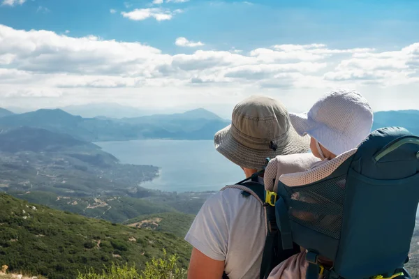 Baba sırt çantasında bebek taşıyor, güzel bir göl, dağ gibi görünüyor. Aile doğada güzel bir yerde yürüyüş yapıyor. Bebek, çocuk konseptiyle tatiller, açık hava aktiviteleri, aktif iyileşme kavramı..