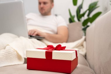 Kırmızı hediye kutusu, kanepeye uzanan, çalışan, dizüstü bilgisayarda, ekoseli evin altında sörf yapan adamın resmi. Noel, sevgililer günü, tatil online alışveriş konsepti..