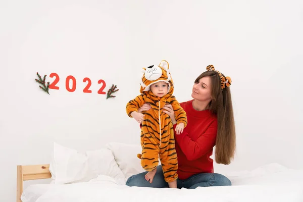 Beyaz tenli esmer kadın, kaplan kostümü giyen, beyaz yatakta oturan, gülümseyen, 2022 numarayı tutan tatlı bir kız çocuğu tutuyor. Doğu Çin takviminde mutlu yıllar kutluyoruz. Boşluğu kopyala..