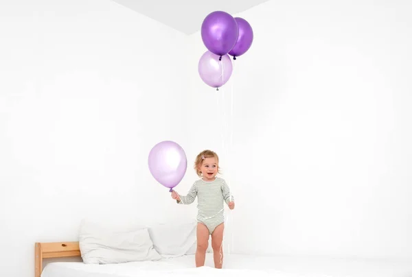 귀엽고 웃긴 금발의 금발의 아기 1, 2 살짜리 아기가 침대에서 놀고 있는 모습입니다. 집에 있는 현대의 파란만장 한 색깔의 커다란 보라색 풍선을 가지고 있습니다. — 스톡 사진