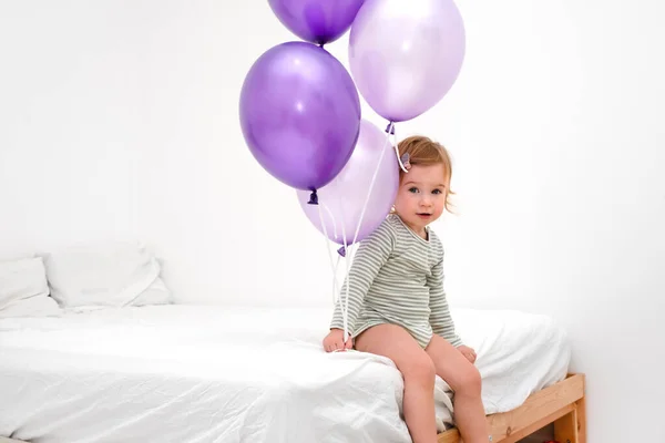 귀엽고 귀엽게 생긴 고양이 금발의 금발의 아기 1, 2 살난 아기가 침대에 앉아 매우 번쩍거리는 색깔의 풍선을 들고 집에서 흰색 배경 카메라를 보고 있다. — 스톡 사진