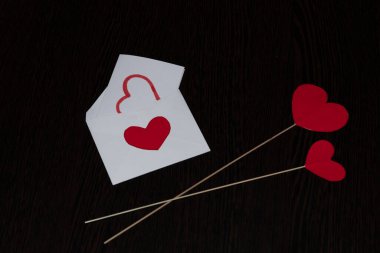 Two red hearts made of cardboard, a white envelope and a message with a painted heart. Attributes of Valentine's day, on a brown, textured background, with a wood texture.